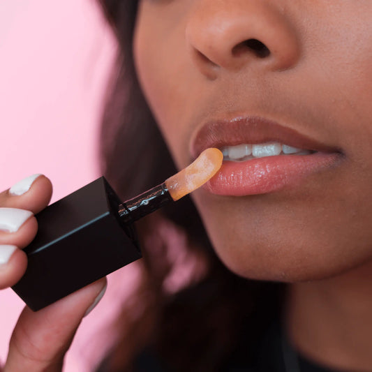 dark skin model applying sheer mandarin lip oil with a pink background