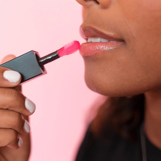 dark skin model applying sheer red lip oil with a blurred pink background
