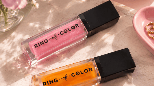 two lip products flat lay next to each other on a table and a small view of a heart shape pink jewerly holder can be seen along with a small flower vase