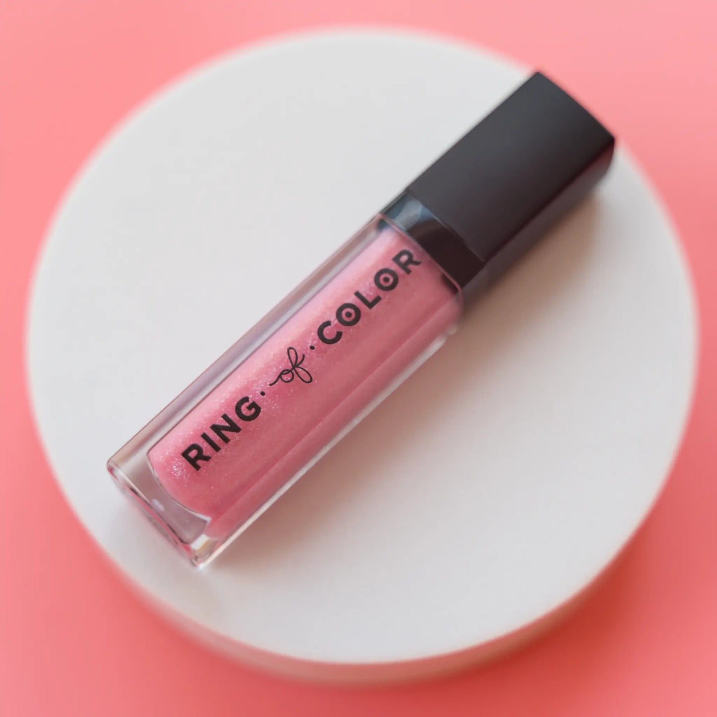 Pink shimmery lip gloss with black cap on a white platform against a pink background
