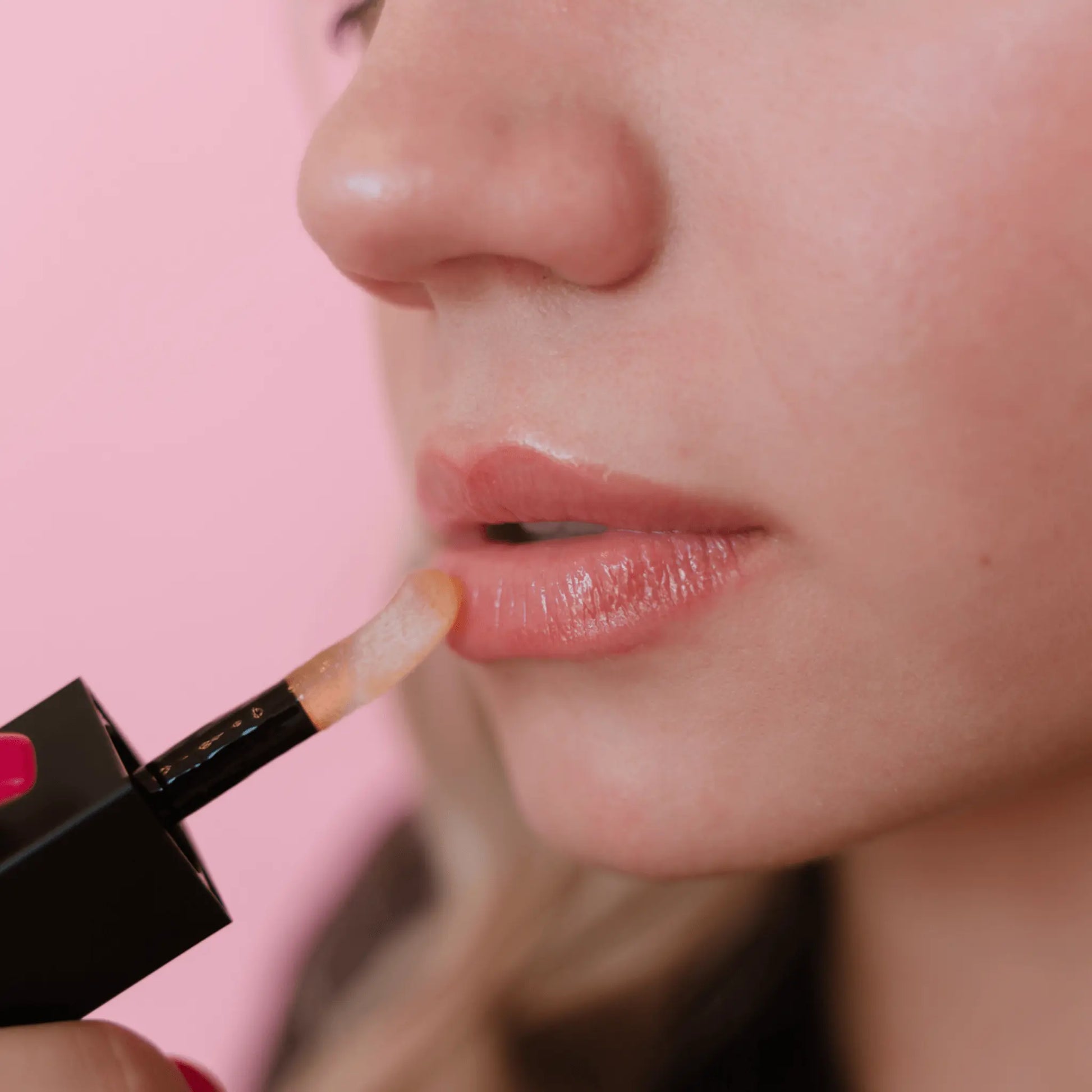light skin model applying sheer mandarin lip oil with a brush on a pink background
