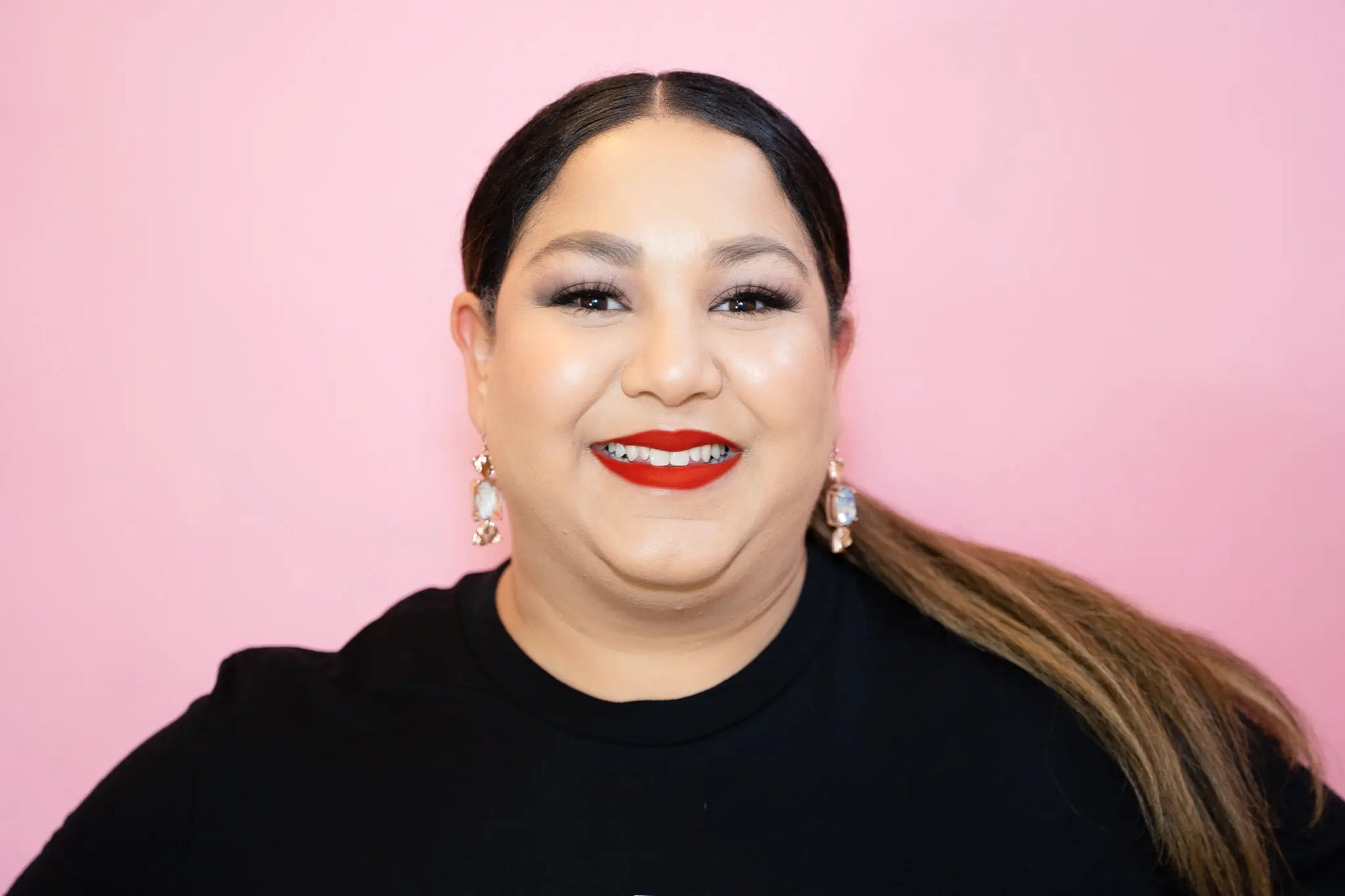 Woman with long hair wearing a black top against a pink background and red lipstick