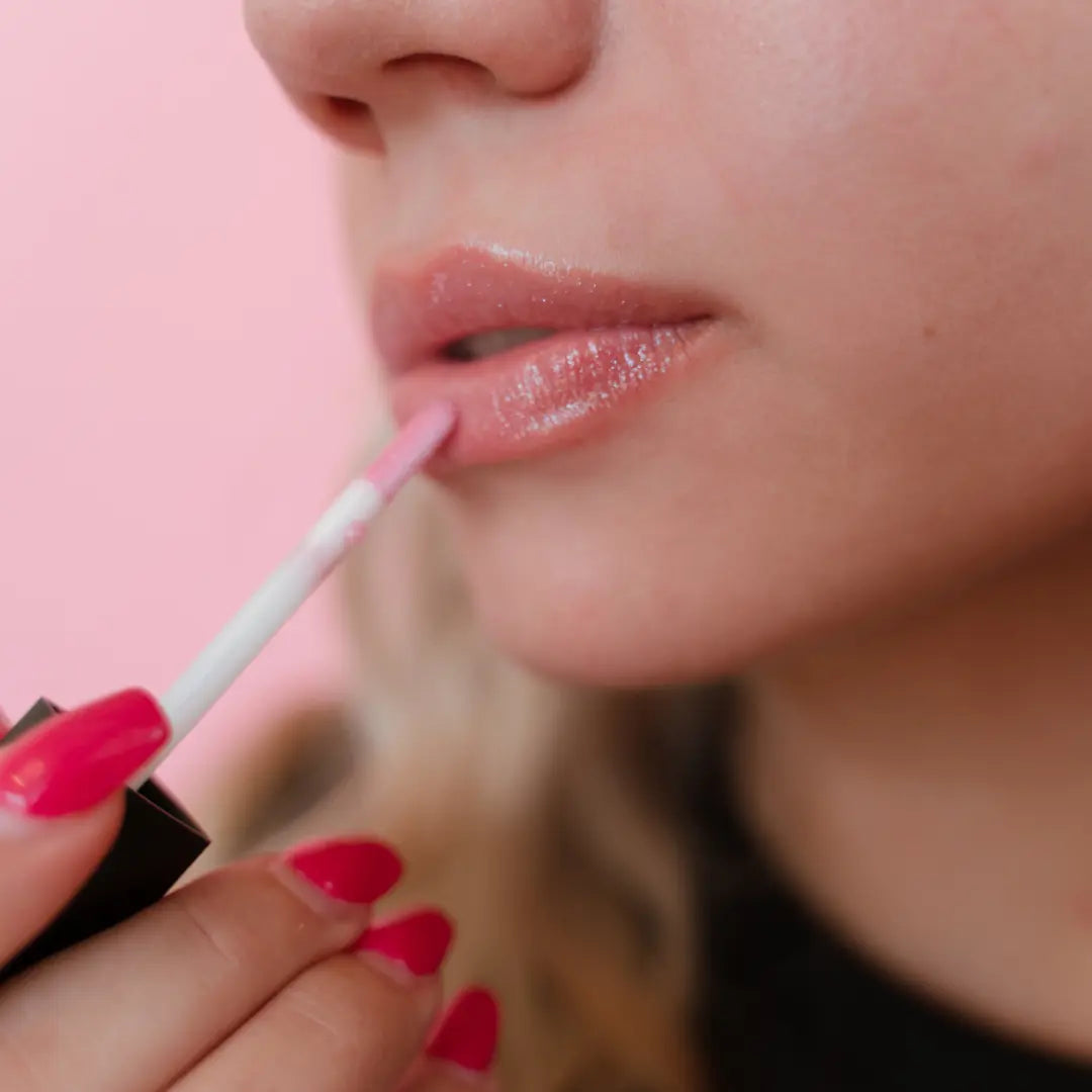 Close-up of a person applying pink shimmery lip gloss with a pink background